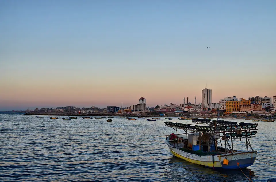 A fishing boat sits in calm water in Gaza port as the sun sets behind the city.